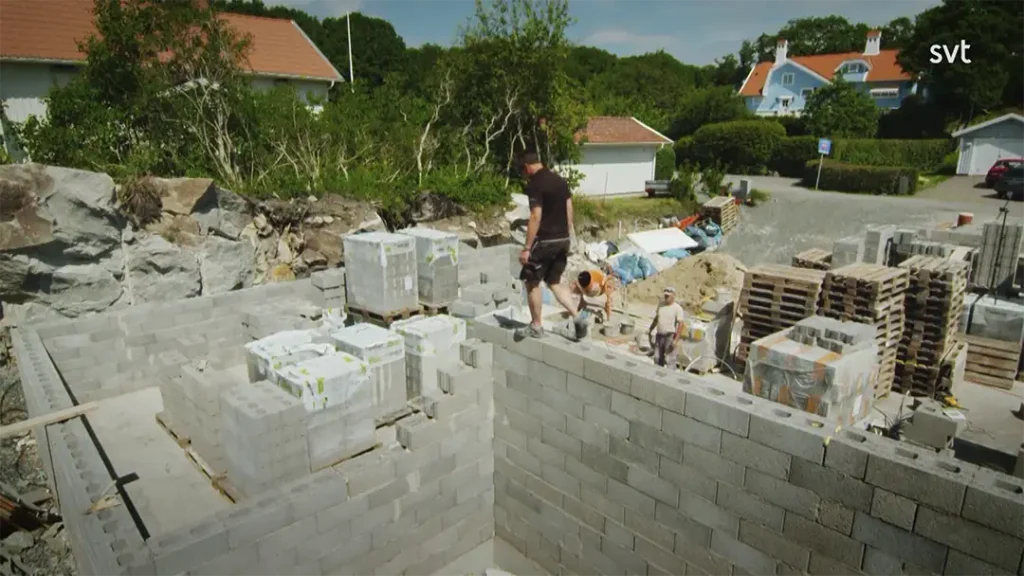 Stiligt - Flera personer arbetar på ett hus under uppbyggnad och staplar gråa betongblock på väggarna, med hem, träd och byggmaterial synliga i bakgrunden.