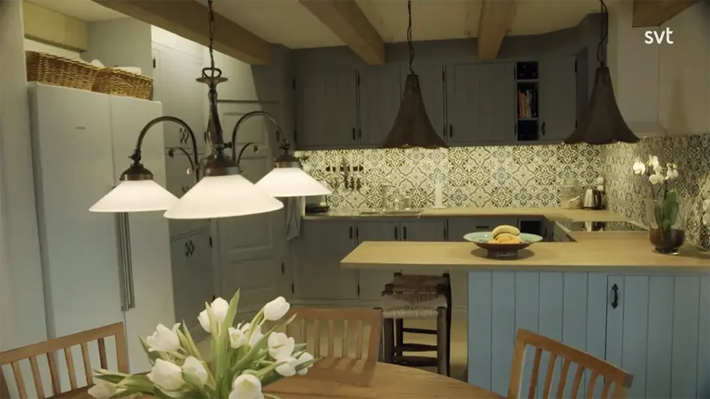 Stiligt - Ett mysigt kök med mönstrat bakstycke, blå skåp, träbjälkar, taklampor och ett matbord med vita tulpaner i en vas.