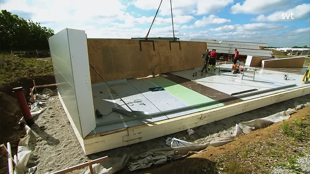 Stiligt - Byggnadsarbetare monterar prefabricerade väggpaneler på en bygggrund på en byggarbetsplats under en delvis molnig himmel.