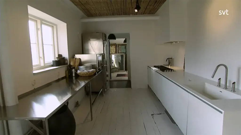 Stiligt - Modernt kök med bänkskiva i rostfritt stål till vänster, vita skåp och diskho till höger, gasspis, samt ett fönster som släpper in dagsljus.