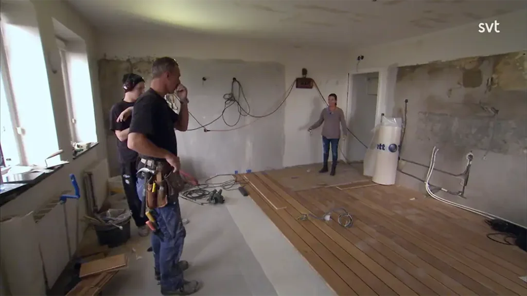 Stiligt - Tre personer står i ett delvis renoverat rum med ofärdiga väggar, synliga ledningar och nya trägolv som installeras.