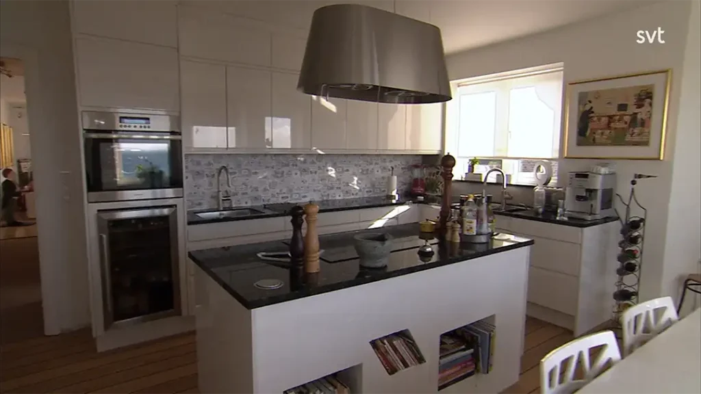 Stiligt - Modernt kök med vita skåp, en central ö med svart bänkskiva, inbyggda vitvaror, en backsplash med mönstrad design och solljus som strömmar genom ett fönster.