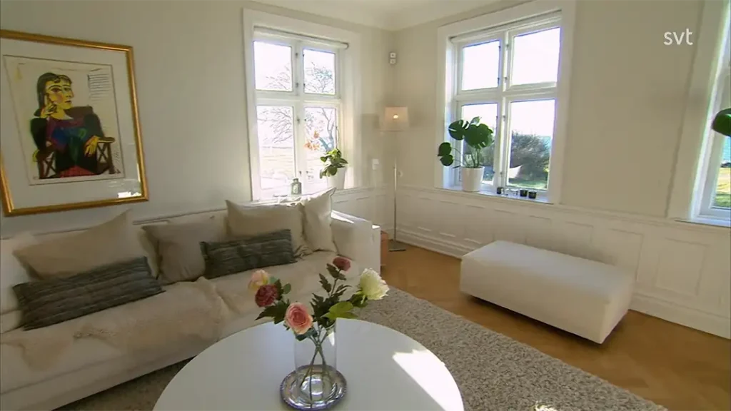 Stiligt - Ljust vardagsrum med vita väggar, stora fönster, en vit soffa med kuddar, en ottoman, ett runt bord med blommor och ett färgglatt porträtt på väggen – perfekt för boende i Borstahusen - Husdrömmar stil.