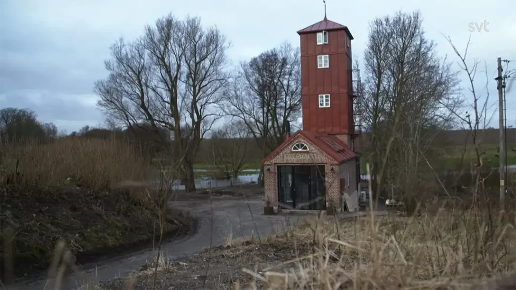 Stiligt - En hög smal röd- och tegelbyggnad med torn står vid en slingrande stig nära en å, omgiven av lövlösa träd och torrt gräs under en molnig himmel — en scen som påminner om Stora Köpinge - Husdrömmar.