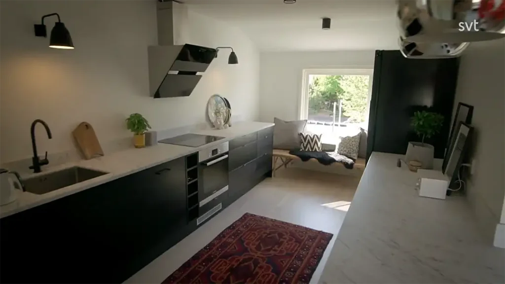 Stiligt - Modernt kök med svarta skåp, vita bänkskivor, en röd matta, väggmonterade lampor och en fönsterplats med kuddar med utsikt över trädgården – ett äkta Mellbystrand - Husdrömmar-inspirerat utrymme.