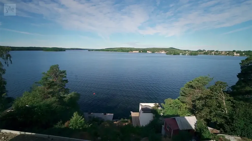 Stiligt - Fridfull utsikt över Mälaren med gröna träd i förgrunden, några byggnader nära stranden och avlägset land under en halvmolnig himmel – perfekt inspiration för Husdrömmar.