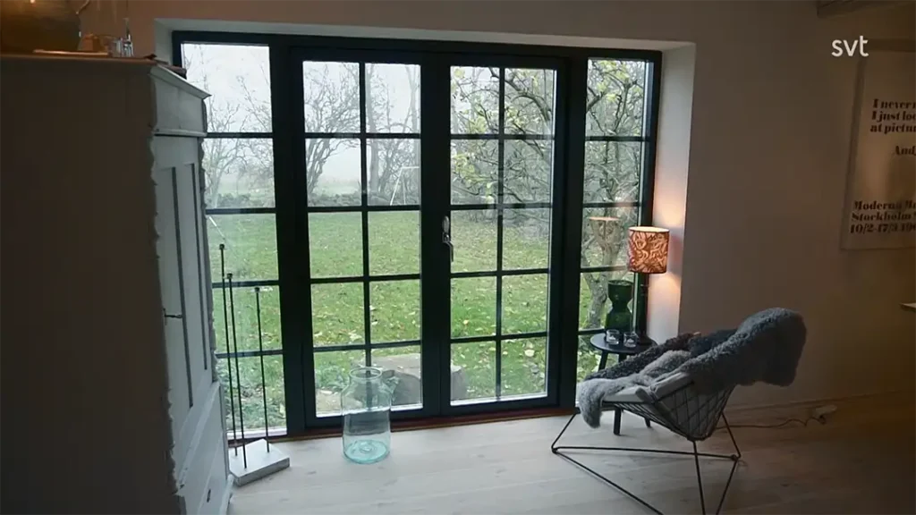 Stiligt - En glasdörr med svarta ramar öppnar sig mot en gräsbevuxen Österlengård med bladlösa träd; inuti skapar en stol med filt, litet bord, lampa och vitt skåp ett mysigt Husdrömmar-inspirerat utrymme.