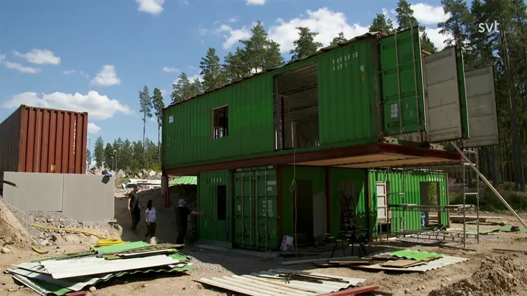 Stiligt - Två gröna fraktcontainrar byggs om till en tvåvåningsbyggnad på en byggarbetsplats i Tranås, omgiven av träd och utspridda material — en sann Husdrömmar i vardande.
