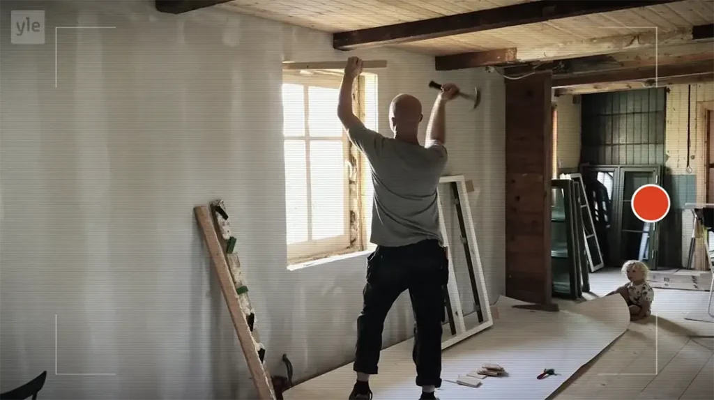 Stiligt - En person använder en hammare för att arbeta på en fönsterkarm i en Kiaby - Husdrömmar renovering; verktyg, ramar och ett barn som sitter på golvet syns.