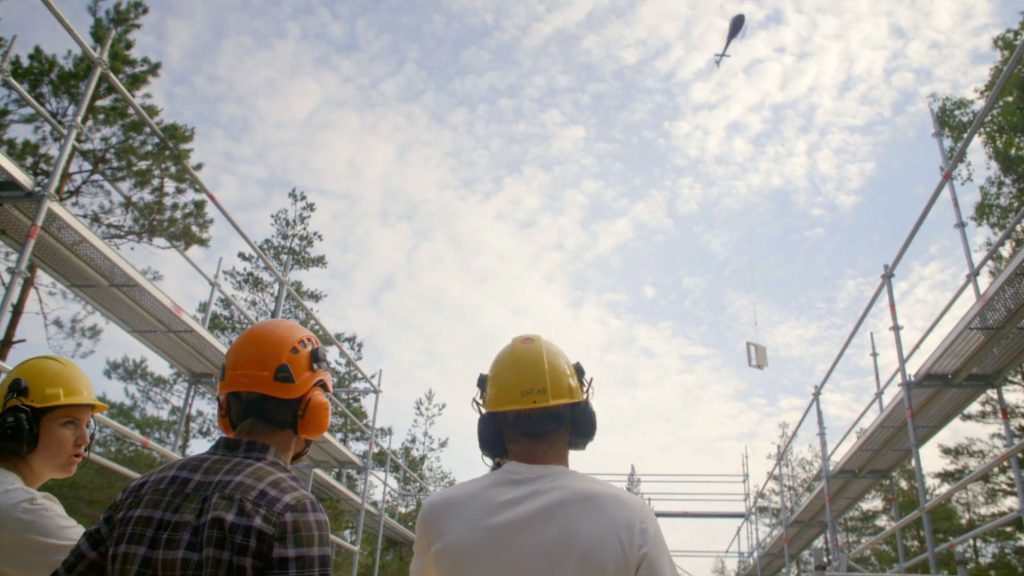 Stiligt - Tre byggnadsarbetare med hjälm och hörselskydd i Huddinge tittar upp när en helikopter transporterar byggmaterial över huvudet, vilket ger en touch av Grand Designs ambition till en arbetsplats omgiven av byggnadsställningar och träd.