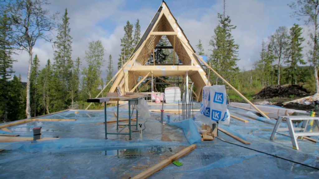 Stiligt - A-ramshus under uppförande på en skogsglänta nära Åre – Grand Designs stil – med synliga träbjälkar, byggmaterial och verktyg utspridda över ett plastklädd golv.