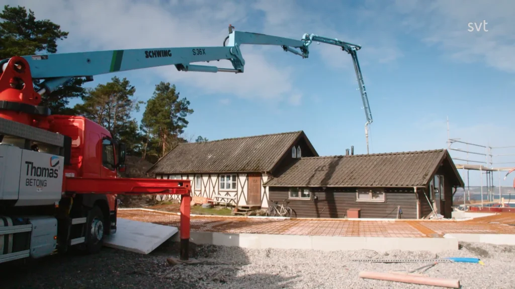 Stiligt - En betongpumpbil häller betong på grunden till en bohuslänsk Husdrömmar byggarbetsplats nära ett hus, omgivet av träd och blå himmel.