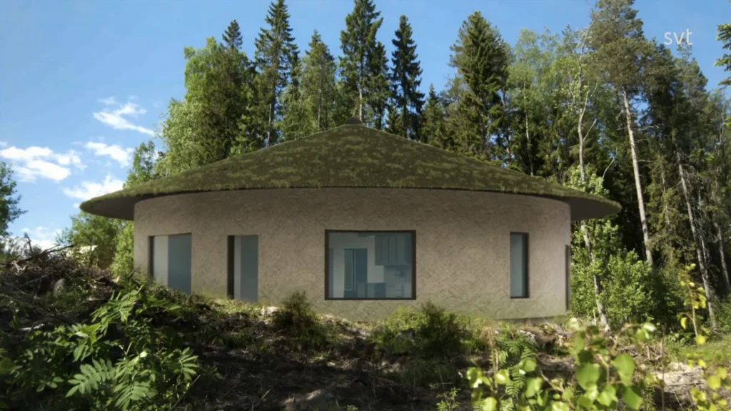 Stiligt - En rund jordtonad byggnad med mossklädd tak står i en skogsglänta nära Kassjö — Husdrömmar inspirerad — omgiven av höga träd och frodig grönska under en blå himmel.