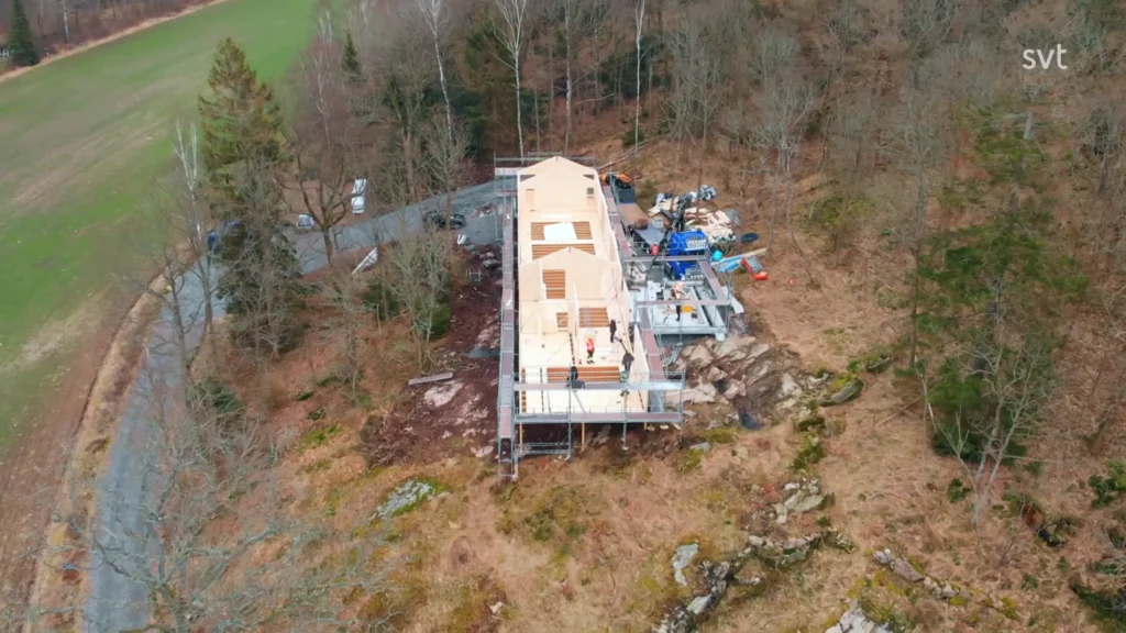 Stiligt - En flygvy över ett rektangulärt Veddige - Husdrömmar hus under uppförande, inbäddat bland träd och utrustning på ett sluttande, delvis röjt skogsområde.