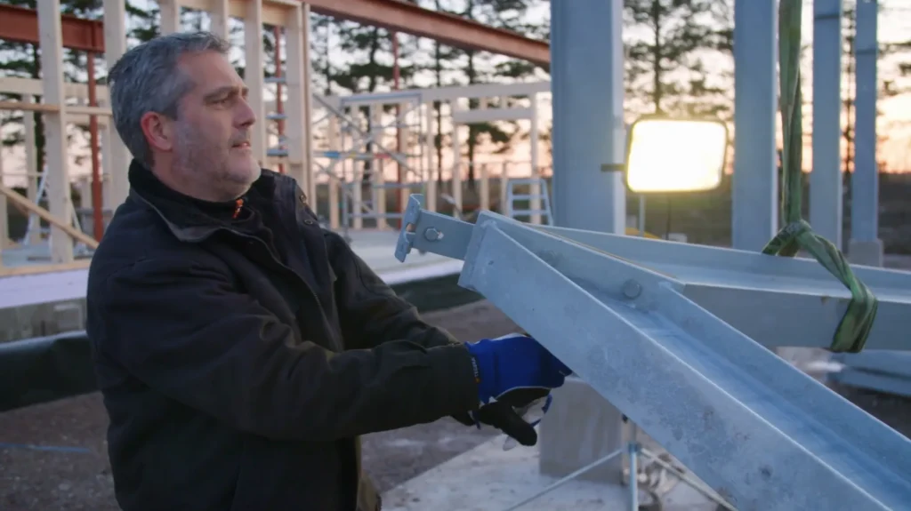 Stiligt - På en byggarbetsplats i Vadstena under solnedgången håller en man i svart jacka och blå handskar en stor metallbalk – en imponerande scen värdig Grand Designs.