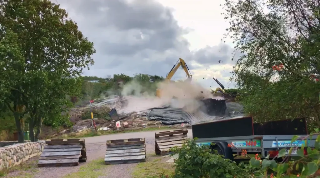 Stiligt - En grävmaskin river en byggnad och skapar ett moln av damm, medan träd och byggbarriärer kantar förgrunden - en stadsförvandling som återspeglar andan i Kattegatt - Grand Designs.