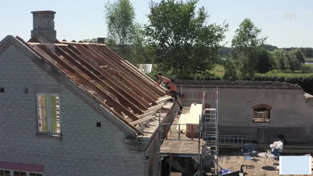 Stiligt - En person arbetar på trätaksstommen till ett tegelhus under renovering i Härnestad – Husdrömmar tar form, med ställningar och byggmaterial runt omkring. Träd och öppna fält utgör den natursköna bakgrunden.
