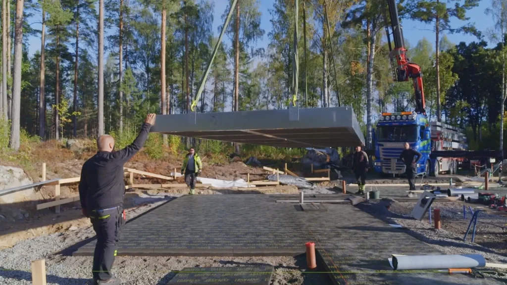 Stiligt - Ett byggteam i Bollnäs guidar en stor prefabricerad betongpanel, sänkt med en kran på en förberedd grund på en byggarbetsplats omgiven av träd, som påminner om Grand Designs projekt.