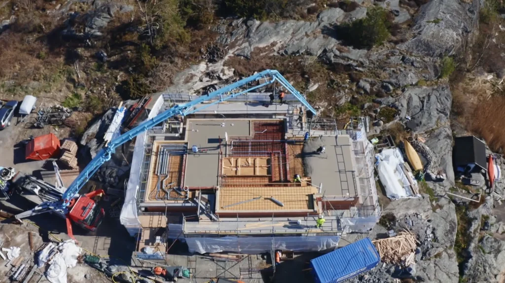 Stiligt - Flygfoto över en Torslanda - Grand Designs-byggnad under uppbyggnad, med arbetare, byggnadsställningar och en stor blå betongpump på plats, mitt i stenig terräng och utspridda byggmaterial.