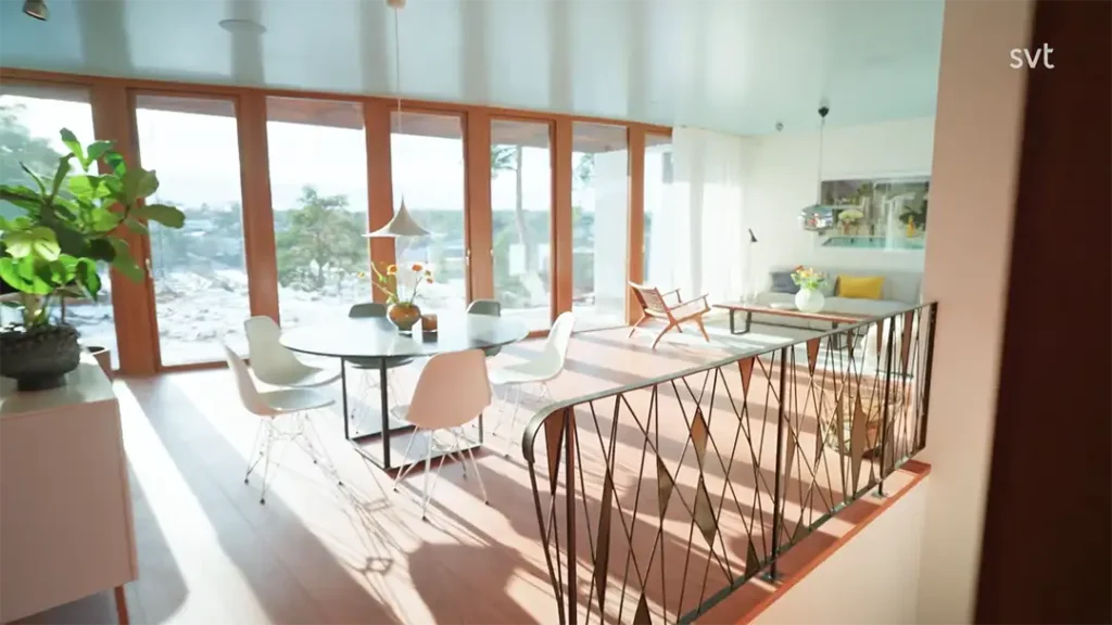 Stiligt - En modern mat- och vardagsrumsdel i Saltsjöbaden - Husdrömmarstil, med stora fönster, ett runt bord med vita stolar, frodiga inomhusväxter och naturligt ljus som strömmar in.