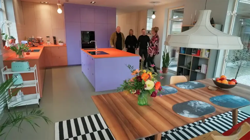 Stiligt - Ett modernt Höllviken - Husdrömmar kök med orange och lila skåp, ett matbord i trä med blommig mittpunkt och fyra personer som står nära köksön.