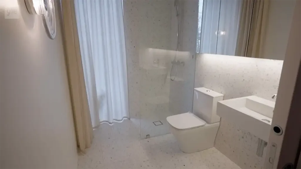 Stiligt - Upplev ett Trångsund - Husdrömmar modernt badrum med inglasad dusch, vit toalett, rektangulärt handfat, stor spegel, samt gardiner från golv till tak vid fönstret.