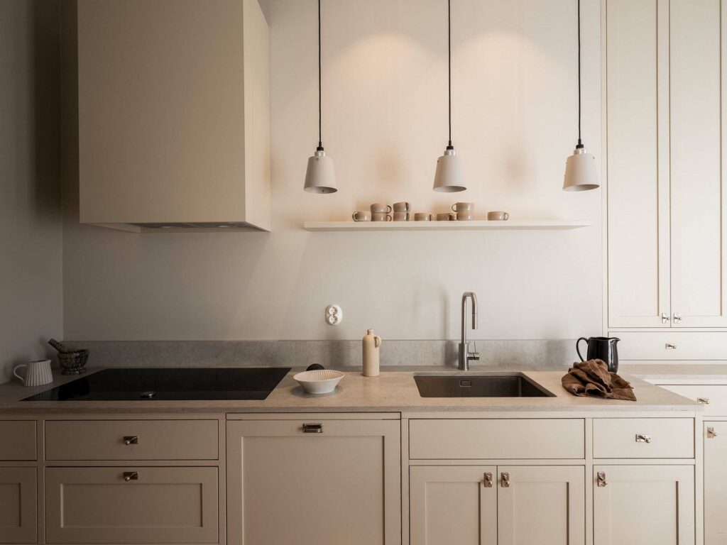 Stylish - The sleek, minimalist kitchen features beige cabinets and a sleek black cooktop. A trio of ceiling lights illuminate the countertop adorned with plates, an electric kettle and a brown towel next to the sink.
