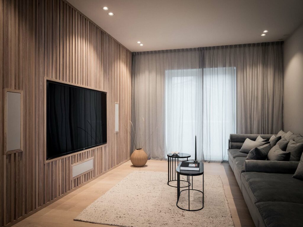 Stylish - A stylish modern living room features a large TV, wood-paneled walls, a gray sofa, and small round tables atop a textured rug. Soft lighting and sheer curtains elegantly cover the window.