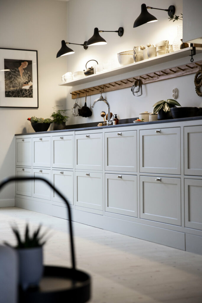 Stylish - Modern kitchen with light gray cabinets, black countertop and stylish wall-mounted shelves. Three ceiling lights illuminate the space, while various interior details add charm. A framed art piece hangs on one wall, reinforcing the room's elegant aesthetic.