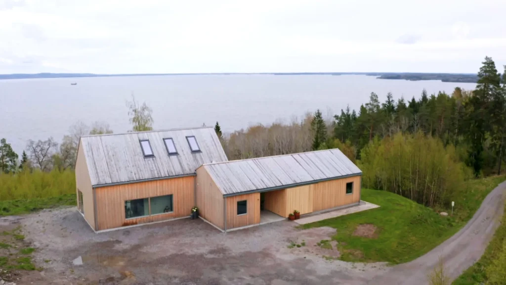 Stiligt - Ett modernt trähus med stora fönster och takfönster står på en grusplats nära en gräsyta i Norrköping, med utsikt över vatten och träd – en sann Grand Designs-inspiration.
