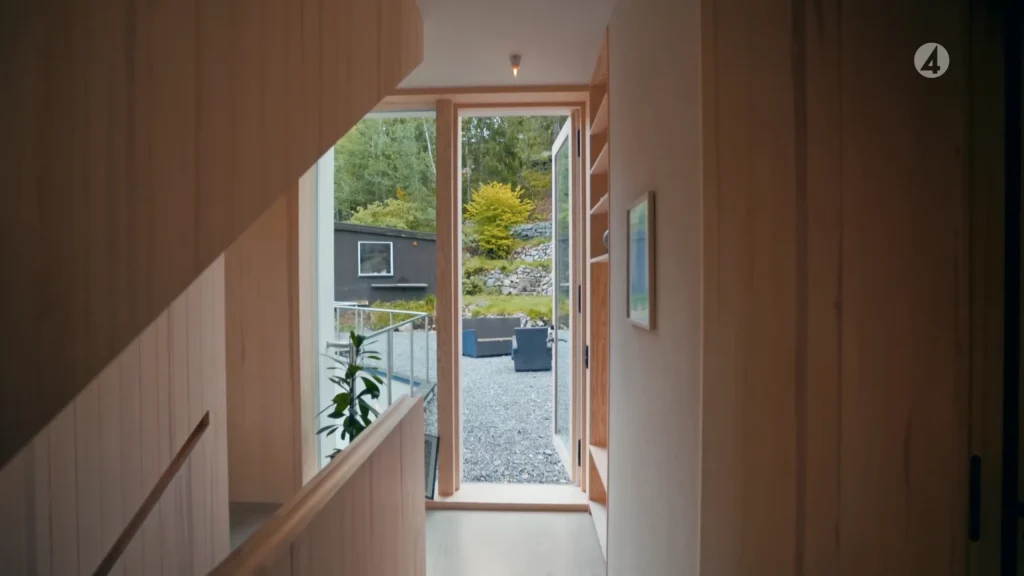 Stiligt - En smal hall med träväggar leder till en öppen glasdörr som avslöjar en Skuru-inspirerad uteplats i grus, uteservering och grönska – som återspeglar den moderna elegansen hos Grand Designs.