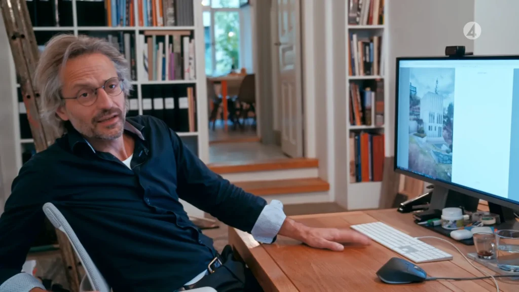 Stiligt - En man sitter vid ett skrivbord på ett hemmakontor och tittar på kameran med en stor bildskärm som visar en arkitektonisk bild från Skuru - Grand Designs. Bokhyllor och en hall syns i bakgrunden.