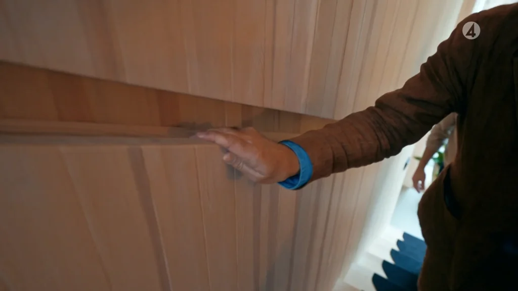Stiligt - En person som bär en brun jacka och blå skjorta öppnar en dold träskåpsdörr bredvid en trappa, och avslöjar smart Skuru-inspirerad förvaring perfekt för alla Grand Designs-entusiaster.