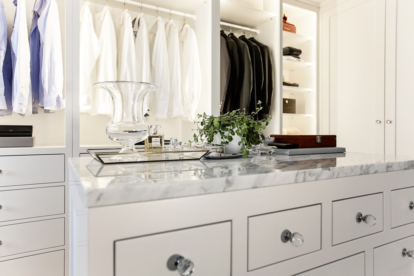 Stiligt - vit walk in closet med snyggt hängda skjortor och jackor, en marmorerad bänkskiva, glasvas, växt och organiserade hyllor med lådor och accessoarer.