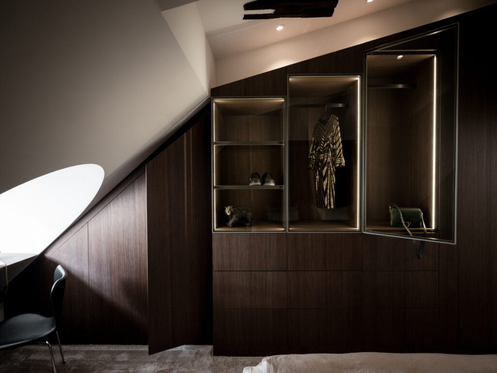 Stiligt - En modern, stilig garderob med träpaneler under ett sluttande tak har en upplyst avdelning med en randig klänning, skor och en handväska.