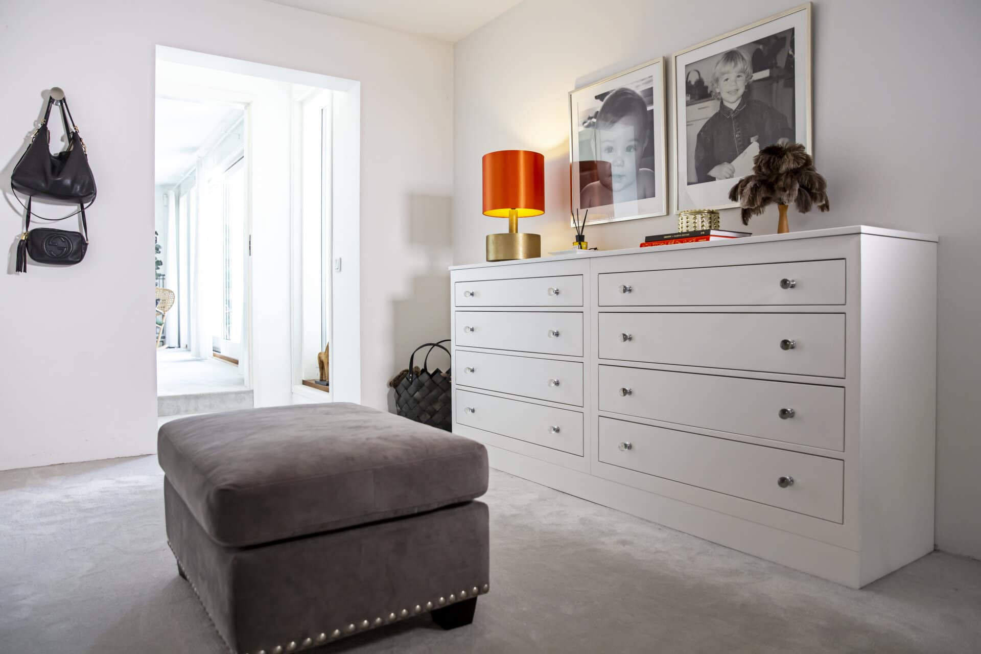 Stiligt - walk in closet i rum som blev över- rummet har en grå ottoman, vit byrå och röd lampa. Svartvita foton pryder väggarna nära entrén i korridoren.