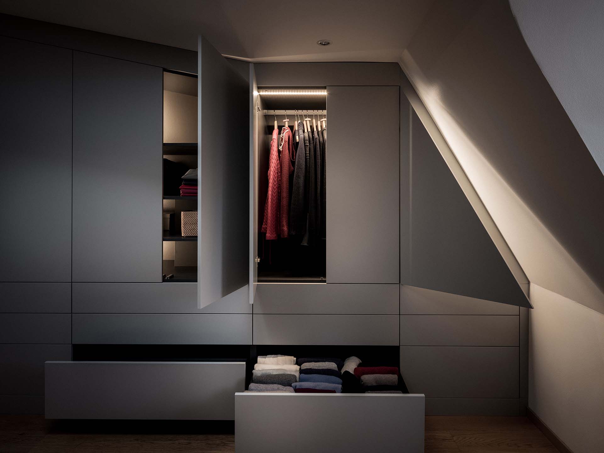 Stylish - Storage slanted ceiling! A modern stylish wardrobe with open doors and drawers shows hanging clothes, folded garments and towels in a dimly lit room.
