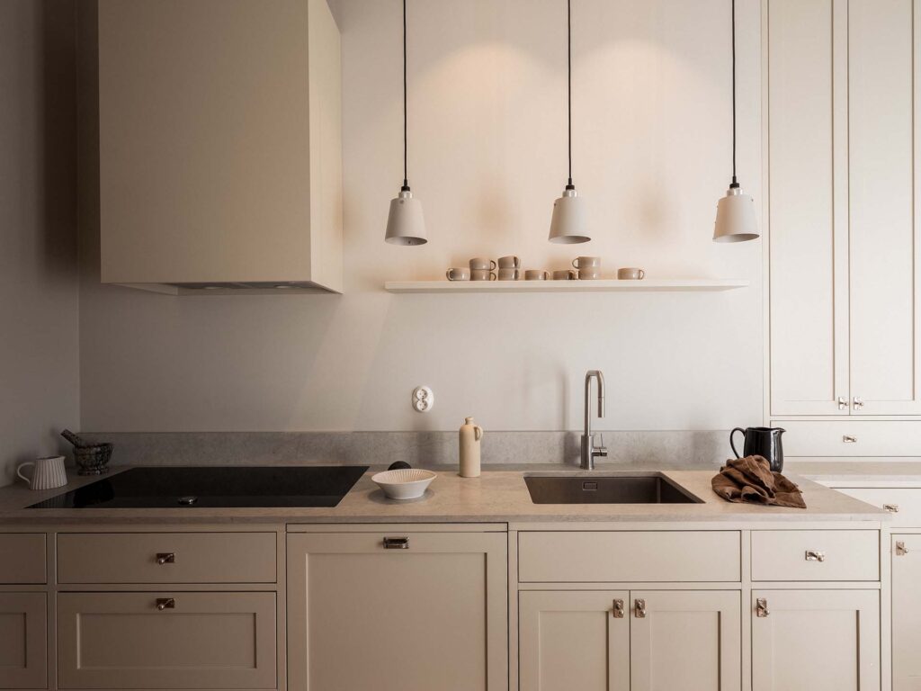 Stylish - A stylish minimalist kitchen features beige cabinets, cooktop, sink and ceiling lights. A shelf displays neatly stacked cups and plates. On the countertop, you'll find a towel and an electric kettle that provide both function and style.