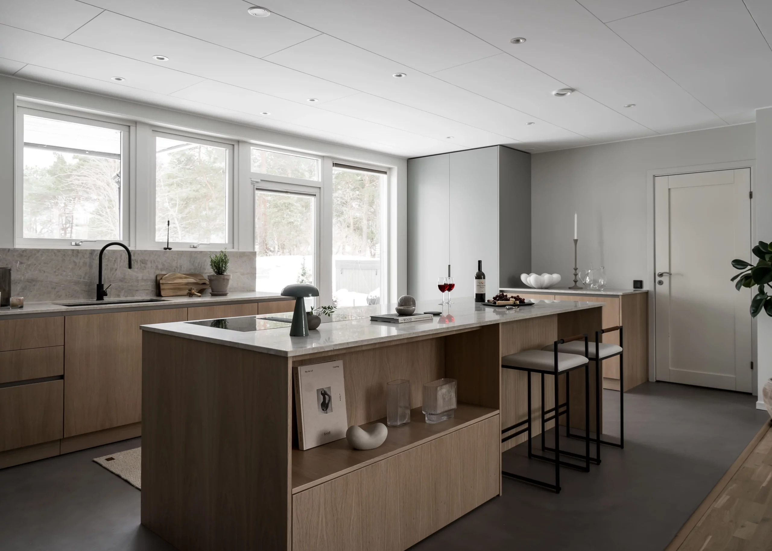 Stylish - Modern kitchen with stylish wooden cabinets, large island and bar stools. A window and a glass door provide natural light. Countertops feature various items, including wine and decor.