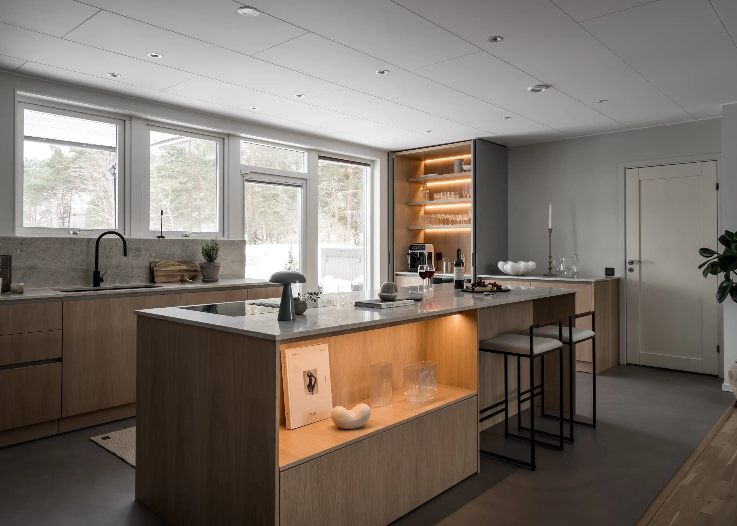 Stylish - Stylish modern kitchen with wood cabinets, large island with bar stools, stainless steel appliances and open shelving. Expansive windows flood the space with natural light, offering a serene view of the snowy landscape.