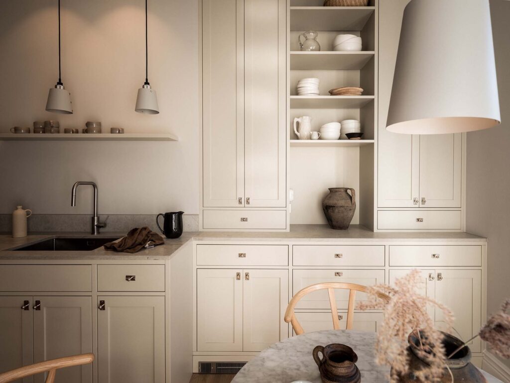 Stylish - A modern kitchen with cream cabinets, a stylish marble countertop, open shelves with porcelain, a sink, a black kettle and pendant lighting over a round table. The color for your site-built kitchen