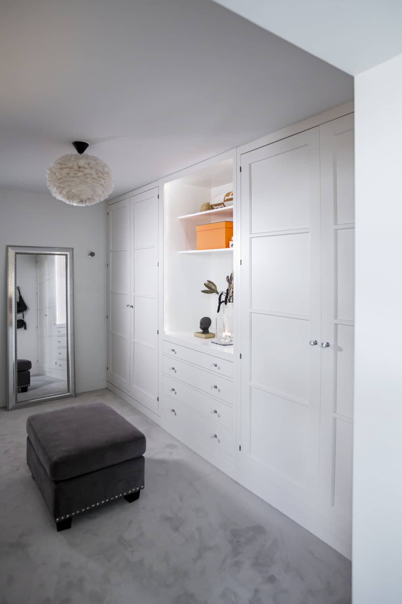 Stiligt - walk in closet i rum som blev över En modern vit walk in closet med stiliga skåp och lådor, en helfigursspegel och en grå ottoman i det heltäckande rummet. En dekorativ lampa hänger elegant i taket.