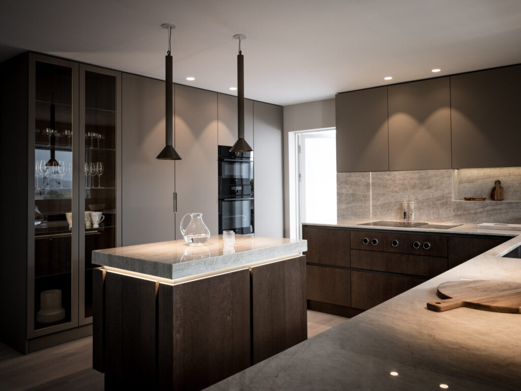 Stiligt - Modernt kök med snygga skåp i mörkt trä, en marmorö med en klar kanna, taklampor och inbyggda apparater. En skärbräda syns på en annan bänkskiva.