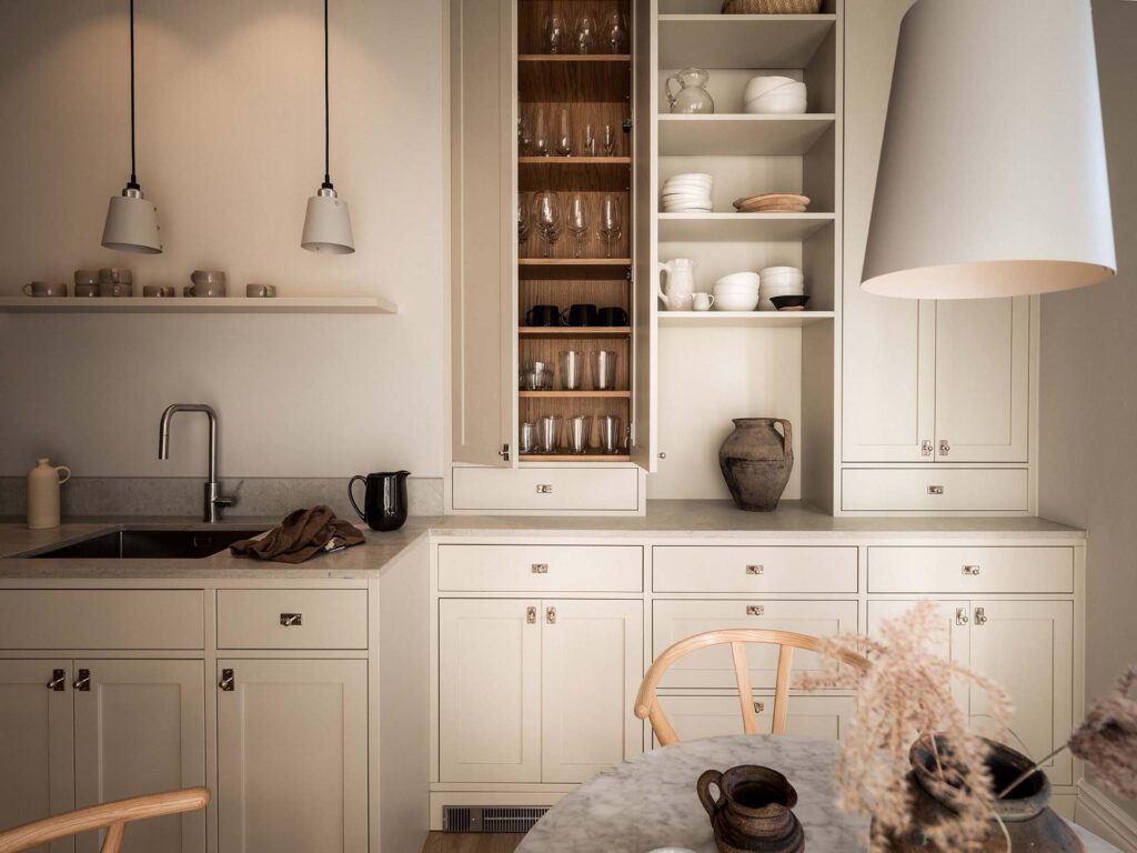 Stylish - A stylish modern kitchen features beige cabinets, open shelves with porcelain and a marble countertop. A sink, an electric kettle and various decorative items add to the chic atmosphere.