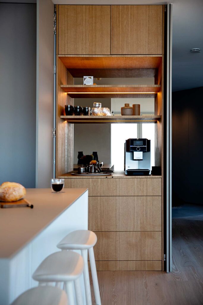 Stylish - Modern stylish kitchen with wooden cabinets, espresso machine and shelves with glasses and mugs. A countertop with three white stools adorns the foreground.