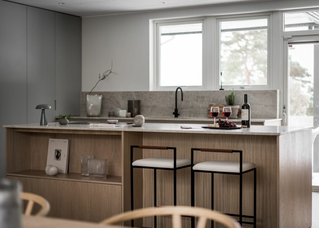 Stylish - A modern stylish kitchen features a light wood island along with two elegant stools and tasteful trinkets. A bottle of wine and two glasses sit enticingly on the counter while sunlight softly filters in through the windows.
