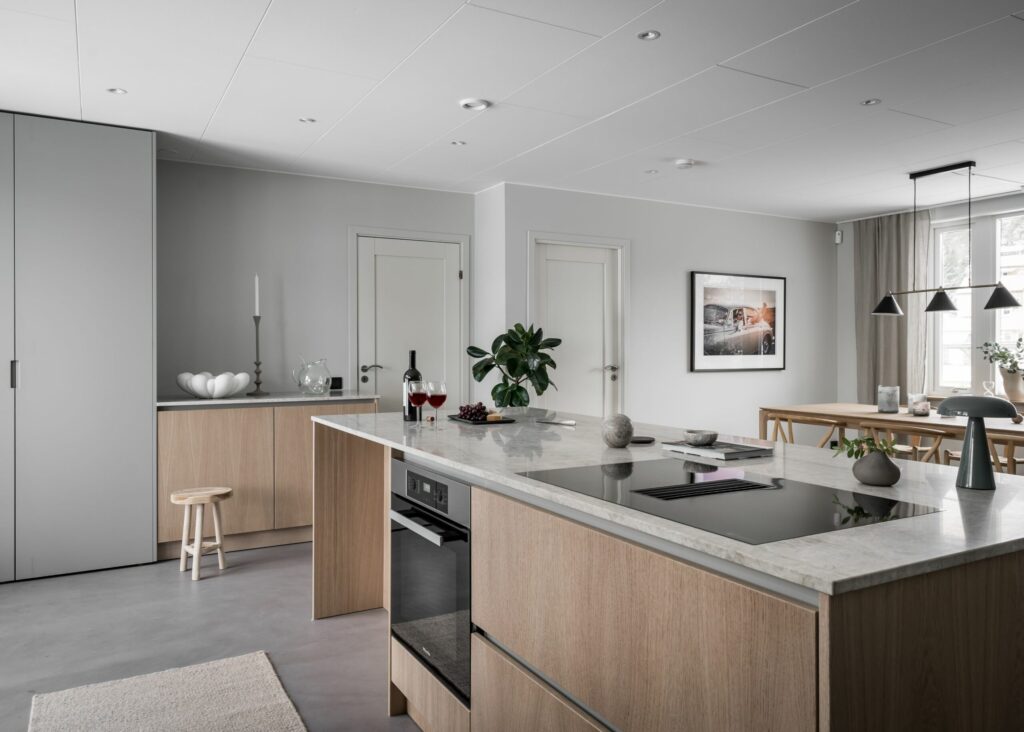 Stylish - A modern kitchen and dining area features stylish design with wooden cabinets, marble countertops, cooktop, wine, fruit bowl and ceiling lights illuminating the dining table. Neutral tones and minimalist decor reinforce the elegant atmosphere.