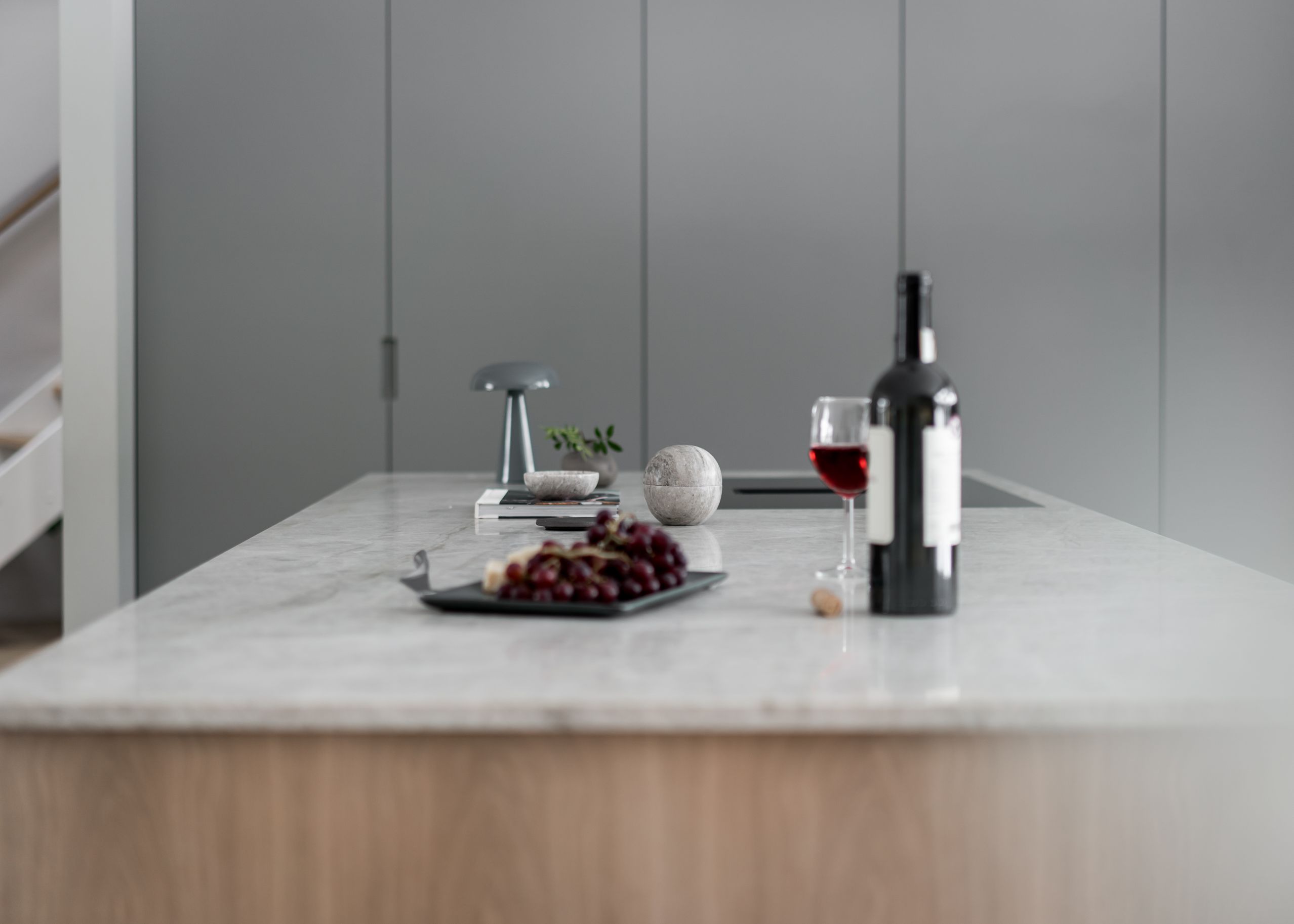 Stylish - A modern kitchen island with a stylish marble countertop showcases a bottle of red wine, a glass, grapes on a plate, a small lamp and decorative items.
