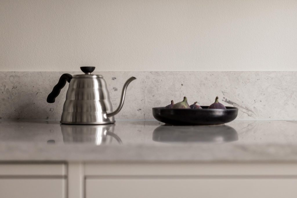 Stylish - A handsome metal kettle sits on a marble countertop next to a black bowl filled with figs.