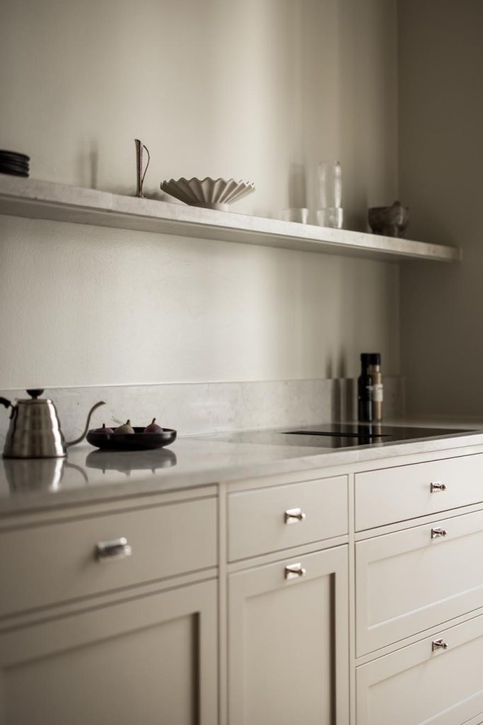 Stylish - The minimalist kitchen features stylish light-colored cabinets, a marble countertop and a matching shelf. A stylish kettle and a plate with cups sit on the counter, while various vases adorn the shelf.